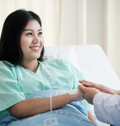 Smiling woman inpatient in a private hospital gown sitting in a bed and holding someone's hand