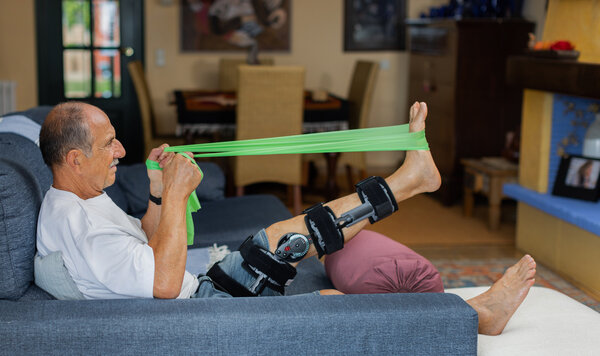 Man wearing a medical brace for his knee sitting on a couch using a resistance band to strengthened his joint muscles ~ HCi health care claims